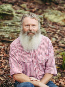 A man sits against a natural rock backdrop looking straight into the camera.